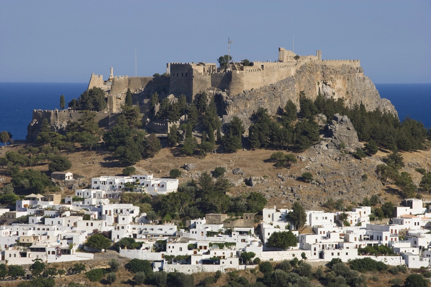 @Image 4 — the backdrop ONLY: the Lindos acropolis above its cascade of white cube houses, allowed into every wide shot as hazed scenery and nothing more.